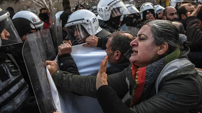 Fotoğraftaki göstericiler polis barikatında durdurulmuş durumda. Kare yakından çekilmiş ve en önde bir kadın polisi itmeye çalışıyor. Arkada da çok sayıda kişi var.