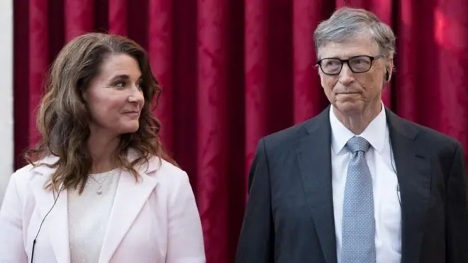 Melinda Gates solda ayakta dururken kafasını sağ tarafındaki Bill Gates'e çevirmiş ve ona bakıyor. Melinda'nın üzerinde parlak beyaz bir üst ve pembe bir ceket varken Bill Gates ise koyu bir ceket, beyaz bir gömlek ve puantiyeli gri bir kravat takmış