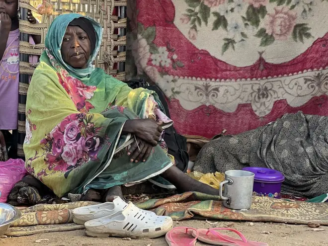 سيدة سوادانية نازحة تفترش الأرض وترتدي الزي السوادني التقليدي الثوب، وبجوارها نعلين.