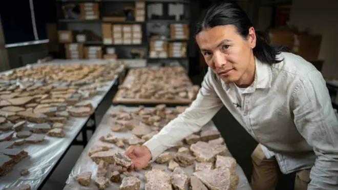 Male archaeologist in a white shirt and beige trousers, with long dark hair, leans over one of three large tables laden with ancient pieces of ancient painted Roman wall. He holds one about the size of a thumb with a blue hue. 