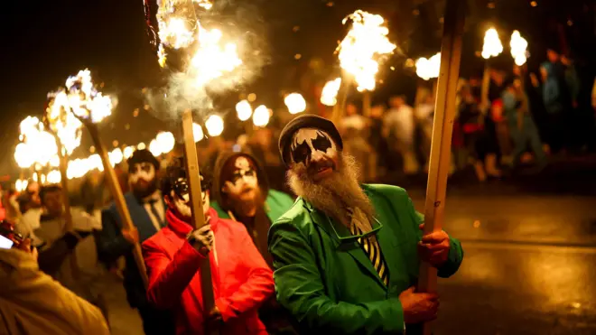 Participants carry fire sticks while wearing masks andf hats and colourful suits during the procession at night.