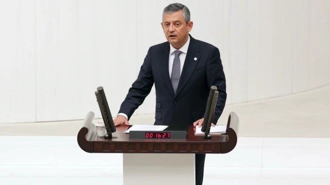 Özgür Özel elini kürsüye koymuş, mecliste konuşma yapıyor. Siyah bir takım giymiş ve karede bir tek o var.