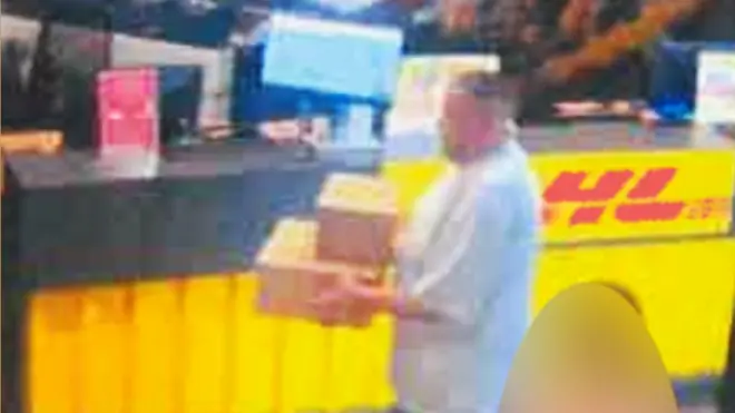 A person stands at a counter holding two cardboard boxes, preparing to ship them. The counter features a bright yellow panel with a red logo, and various signs and equipment are visible on the countertop in the background.