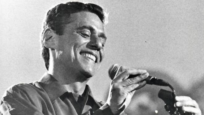 Foto em preto e branco mostra Chico Buarque sorrindo e tirando microfone do pedestal