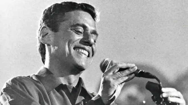 Fotobrabet jogo do pinguimpreto e branco mostra Chico Buarque sorrindo e tirando microfone do pedestal