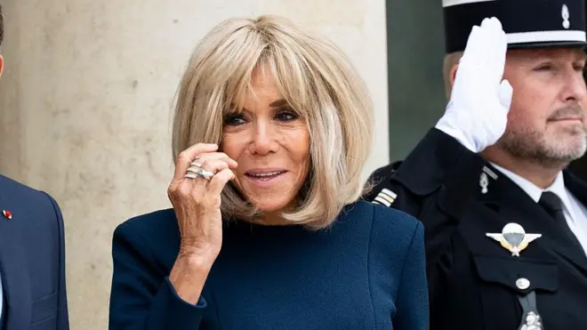 Brigitte Macron a une épaisse chevelure blonde coiffée au carré avec une frange et porte une robe bleue à manches longues. Un salut officiel est entendu en arrière-plan.