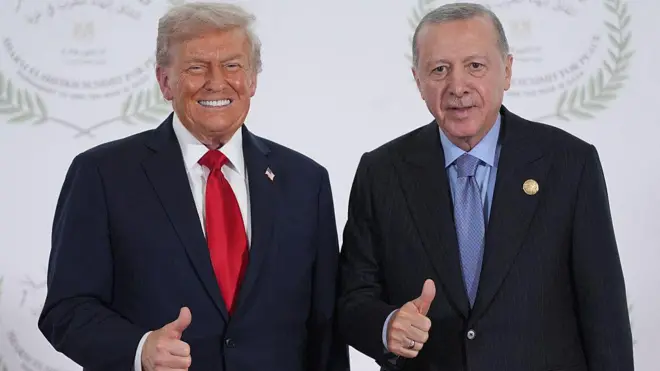 Trump solda Erdoğan sağda, siyah takım elbiseler giyinmişler, elleriyle baş parmaklarını kaldırıp, thumbs up, beğeni işaret yaparak kameraya gülümsüyorlar. Arka planda beyaz bir fon var. 