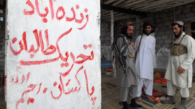 سه مرد با لباس محلی پاکستانی ایستاده‌اند که یکی از آنها سلاح ای‌کی روی دوشش دارد و یکی دیگر جلیقه نظامی پوشیده است