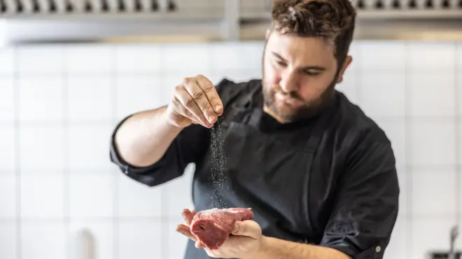 Muškarac u crnom soli komad mesa pre kuhanja