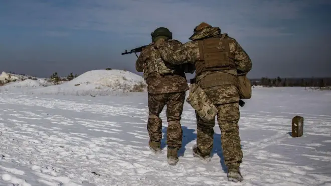 Двоє військових у зимовій формі й бронежилетах йдуть по засніженому полю. Вони зображені зі спини. Один тримає автомат, а другий підтримує його за бронежилет, ніби допомагає йти. На землі лежить товстий шар снігу, а вдалині видно низькі пагорби та дерева. Небо ясне, з легкими хмарами, світло денне.