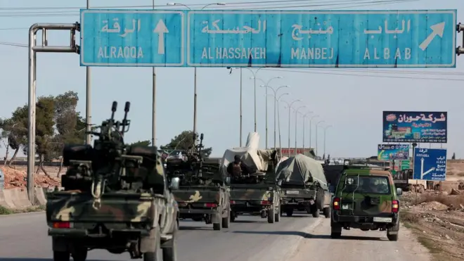 Otobanda ilerleyen silahlı askeri kamuflaja boyanmış arazi araçları içlerindeki askerler arkadan görünüyor. Otobanda Alraaqqa, Al Hassakeh, Manbej ve Albab yönlerini gösteren bir trafik işareti var. 