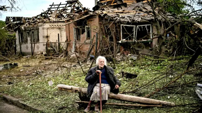 A woman sits on a fallen log in front of two houses that have been demolished