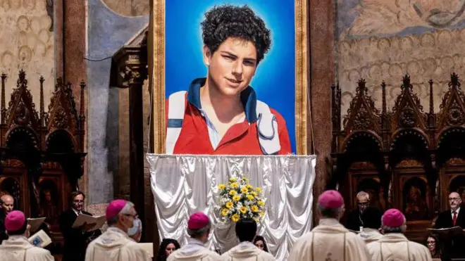 Un tapiz de Carlo Acutis, un adolescente, y personas con atuendos religiosos mirando.