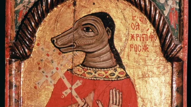 A imagem mostra um ícone religioso antigo de São Cristóvão representado com corpo humano e cabeça semelhante à de um cão. A figura veste uma túnica vermelha ornamentada, segura uma cruz na mão e aparece diante de um fundo dourado com halo e inscrições em caracteres gregos, em uma pintura de estilo bizantino marcada pelo envelhecimento e por uma rachadura vertical no painel.