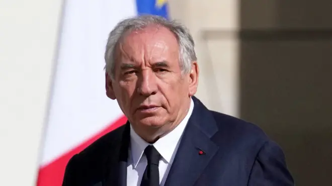 Prime Minister François Bayrou dressed formally in a dark suit, white shirt, and dark tie. He has grey hair and serious expression on his face. He’s standing outdoors, and behind him are two partially visible flags — one appears to be the French tricolor (red, white, and blue), and the other is the European Union flag. There is a small red pin on his left lape