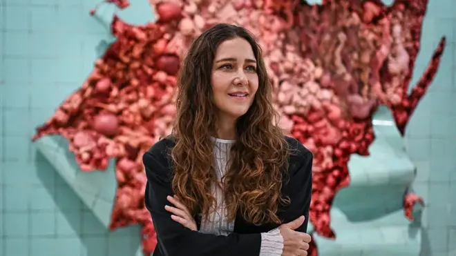 Adriana Varejão, uma mulher de cabelos longos e ondulados, posa de braços cruzados diante de uma instalação artística que remete a carne e vísceras expostas, em tons intensos de vermelho e rosa. Ao fundo, azulejos claros contrastam com a obra orgânica e dramática, criando uma imagem que mistura delicadeza e impacto visual.