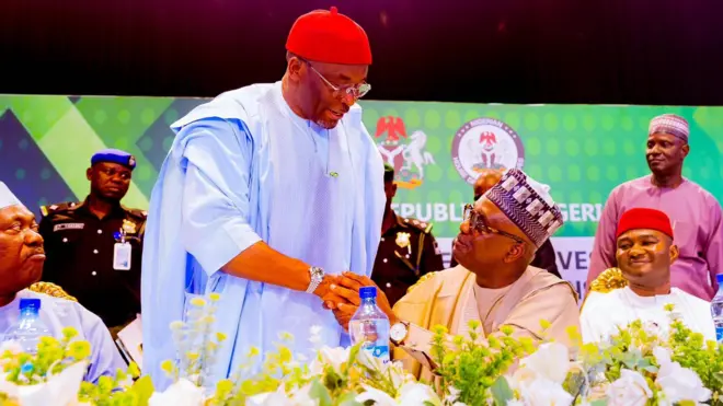 One man stand up wearing red cap shaking anoda man wey sit down, police officer dey di back