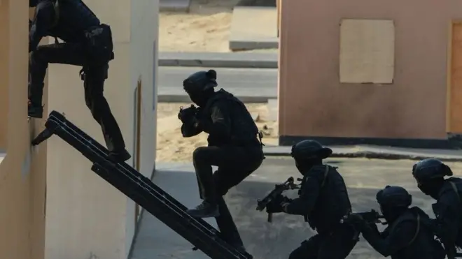 Heavily armed security agents dressed in black scramble up a stepladder to enter the first floor window of a building
