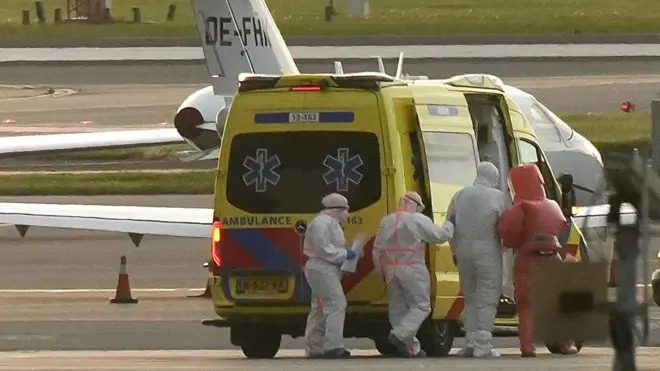 Equipos sanitarios trasladan a una persona con equipo de protección a una ambulancia en la pista de un aeropuerto