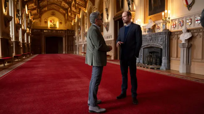 Prince William showing Eugene Levy inside Windsor Castle, they are in a large room or corridor with red carpet and ornate furnishings