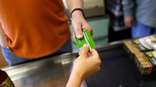 Bir market kasası önünde alışveriş yapan bir erkek, ödeme için yeşil renkli bir kredi kartını uzatıyor.