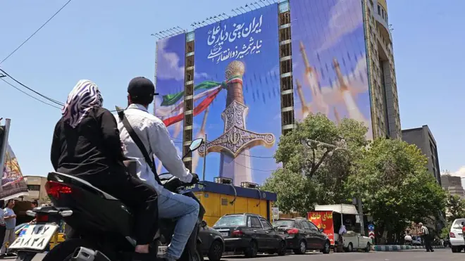 تہران میں موٹر سائیکل پر سوار دو افراد ایک عمارت پر بنے بڑے دیوارگیر نقش کے پاس سے گزر رہے ہیں جس میں خنجر اور داغے گئے میزائل دکھائے گئے ہیں