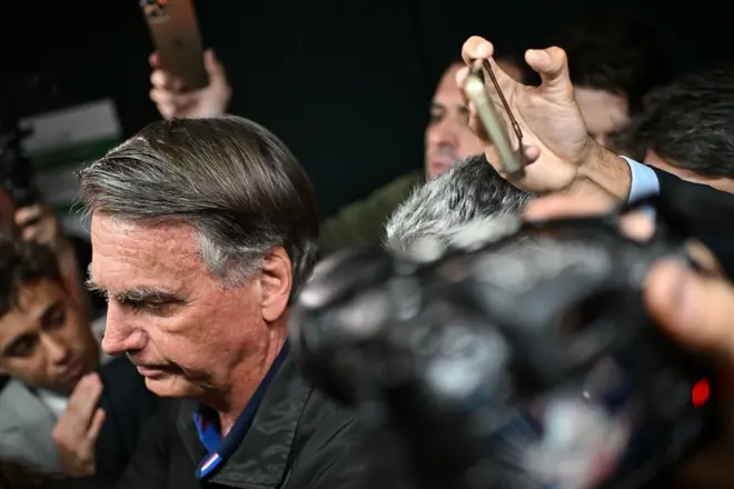 Bolsonaro olhando para a frente com olhar sério, rodeado por jornalistas