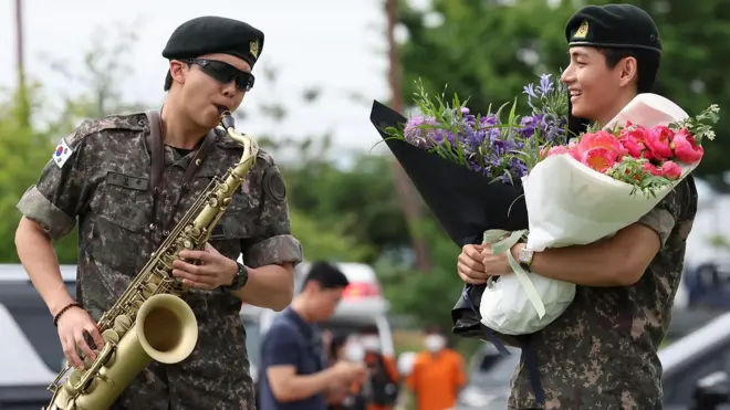 RM, Kim Namjoon is wearing his military uniform and sunglasses, he is playing the saxophone. V, Kim Tae-hyung, is looking at RM, also clad in military uniform, holding two bouquets of purple and pink flowers. 