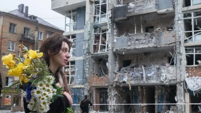 一名手捧花束的女子于 2025 年 5 月 25 日走过基辅谢甫琴科区的一栋高层住宅大楼。她的头发部分散落在额头上。街道上,她身后是一栋遭俄罗斯无人机袭击而严重受损的多层建筑物。