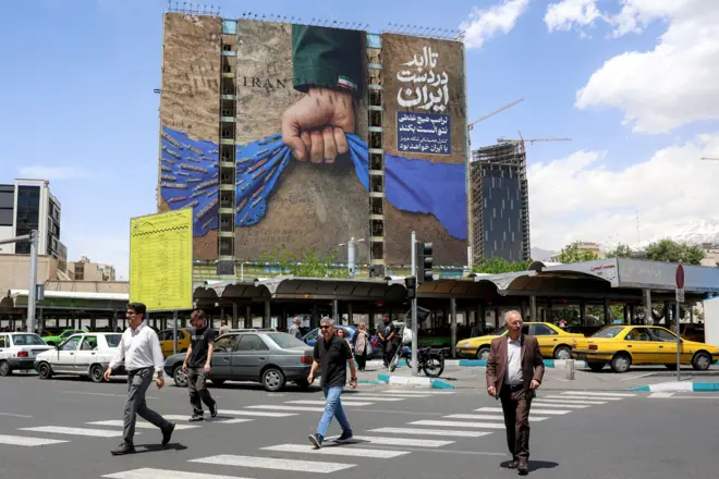 أشخاص يعبرون شارعاً في ساحة في طهران وتظهر لوحة إعلانية على واجهة مبنى تصور مضيق هرمز مع عبارة باللغة الفارسية تقول "إلى الأبد في يد إيران"، في 6 مايو/أيار 2026.