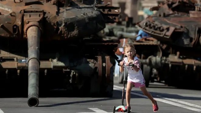 Devojčica vozi trotinet ispred hrpe uništenih tenkova