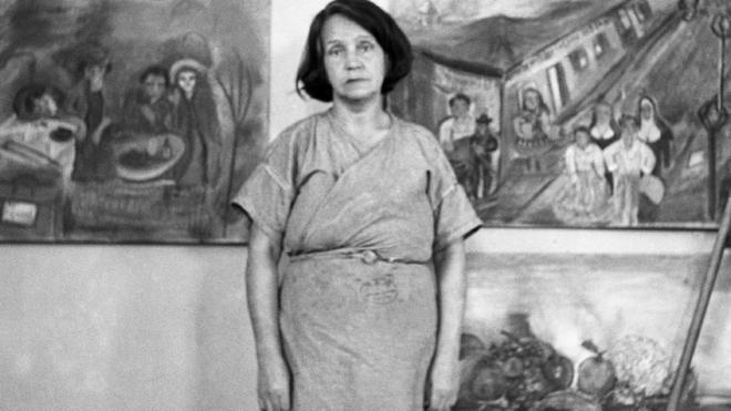Fotografia em preto e branco mostra mulher com jaleco em frente a diversos quadros