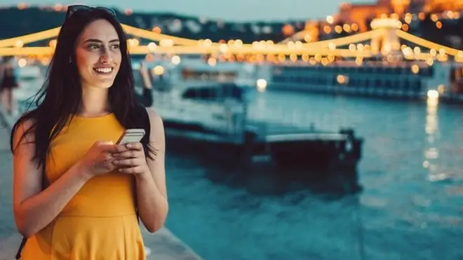 Young woman with a phone in her hand. She's smiling, wearing a yellow dress and walking by the river in Budapest at dusk.