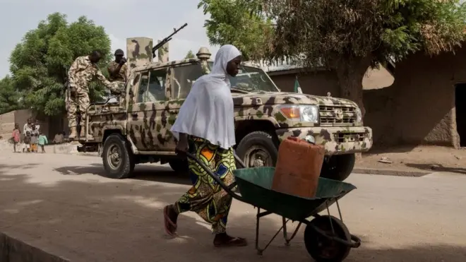 A woman pushes a wheelbarrow carrying a jerrycan filled with water as Nigerian soldiers patrol in the town of Banki in northeastern Nigeria on April 26, 2017