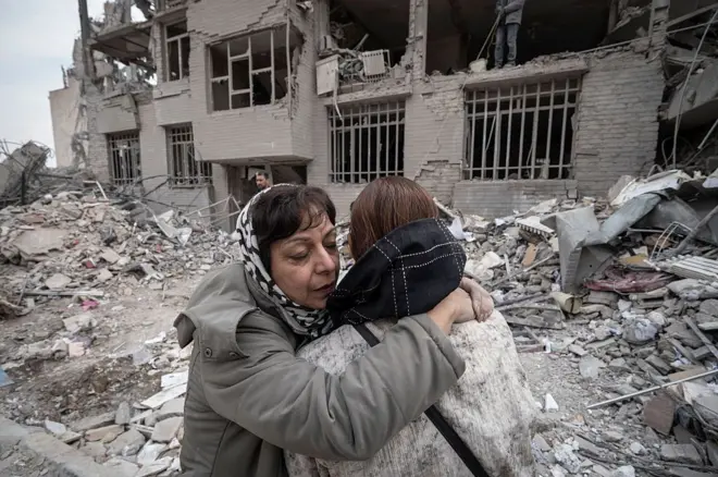 دو زن در آغوش هم در حالیکه پشت سرشان خانه‌ای ویران شده قرار دارد