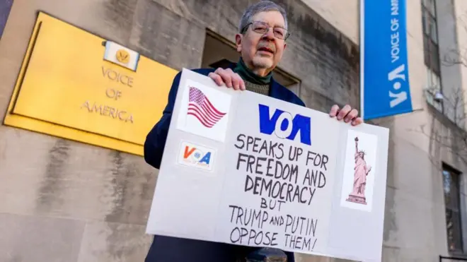 Steve Lodge, whose father Robert Lodge was a correspondent at VOA, stands in protest in front of the organisation's headquarters in Washington DC. Dressed in a navy coat, he holds a banner that reads "VOA speaks up for freedom and democracy, but Trump and Putin oppose them!"