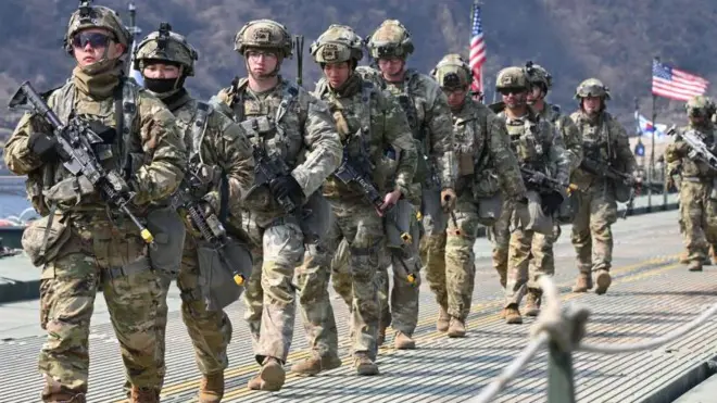 Des soldats américains forment une colonne. Derrière eux  flotte le drapeau américain