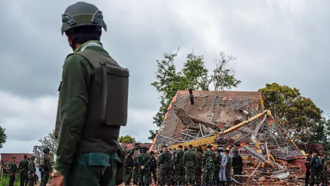 A Cambodian soldier stands on guard near a destroyed building at the border between Thailand and Cambodia. He watches a team of soldiers and officials inspecting the site.   