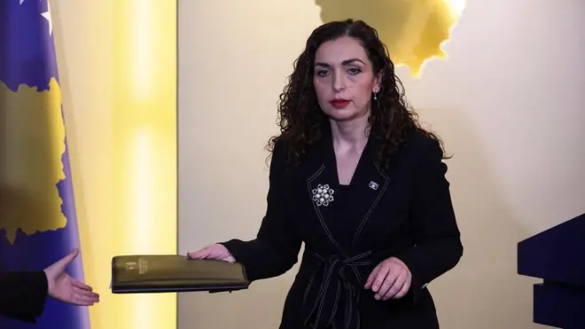 Војса Османи гледа у под у десној руци држи свеску, а десно од ње је застава Косова