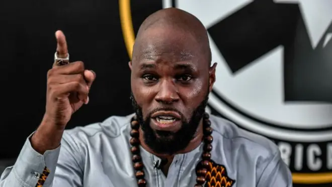 Kemi Seba, vêtu d'une chemise grise, est de face. Il porte un collier de perles en bois et son bras gauche est levé, l'index pointé vers le haut. À l'arrière-plan, on aperçoit partiellement le logo de l'ONG Pan-Africanist Emergency.
