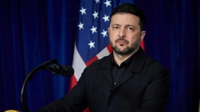青いカーテンと星条旗を背にしたゼレンスキー氏。黒い上着とシャツを着て、厳しい表情をしている