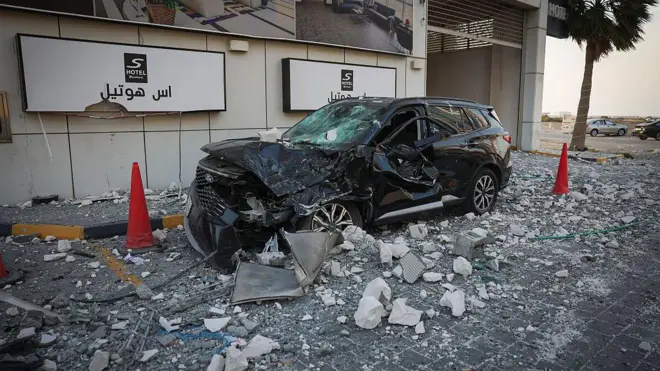 A wrecked black car sits between two red traffic cones among debris outside a hotel in Manama, Bahrain on Sunday 1 March 2026, with two other cars behind a palm tree in the background.