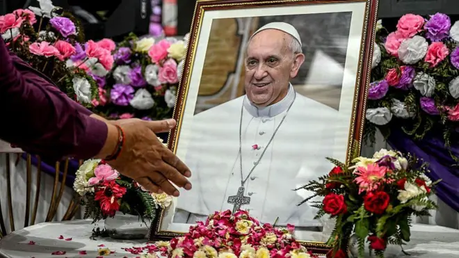 Руки пожилой прихожанки сложены в молитве у портрета папы Франциска в окружении цветов