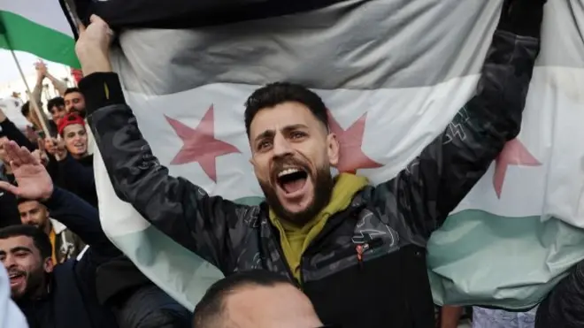 Un hombre entre una multitud sostiene una bandera siria sobre su cabeza en señal de celebración.