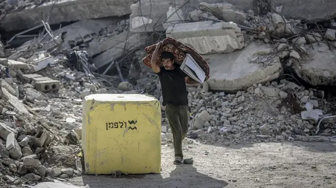 رجل يحمل مرتبة مطوية في غزة يمر بجوار كتلة خرسانية صفراء وضعها الجيش الإسرائيلي، وتظهر الأنقاض في الخلفية