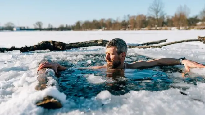 Um homem de cabelos grisalhos e barba toma banho em lago congelado durante o inverno