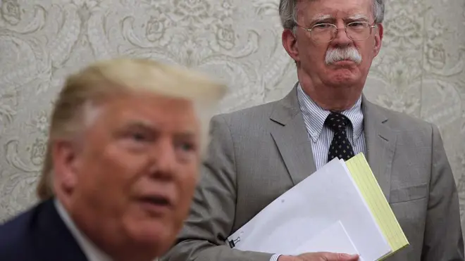 John Bolton, hold documents and yellow legal pad, stand behind Donald Trump, wy sidon for Oval Office. 