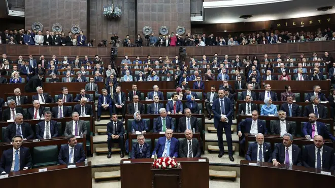 29 Nisan'da Meclisteki AKP grup toplantısında bir araya gelmiş, çoğunluğu erkeklerden oluşan partili milletvekilleri koyu renk takım elbiseleriyle koltuklarda oturmuş ve ön sıradaki Cumhurbaşkanı ve AKP Genel Başkanı Recep Tayyip Erdoğan'ın önündeki masada kırmızı ve beyaz renkli çiçekler var, arkadaki balkon kısmında izleyiciler ayakta duruyor