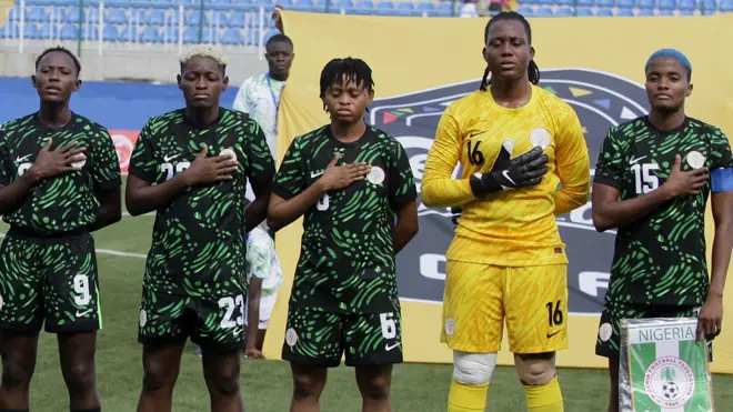 Five Nigeria players, including a goalkeeper wearing all yellow and a captain holding a Nigeria pendant, stand with their left hands on their chests to mark a national anthem while lining up before a match