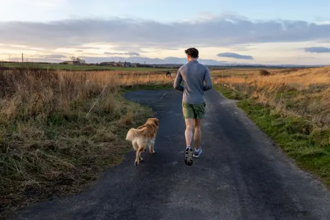 Um homem jovem adulto correndo com seu cachorro por uma estrada rural. Ele veste roupas esportivas, segura a guia do cão, e o sol se põe sobre a paisagem ao redor